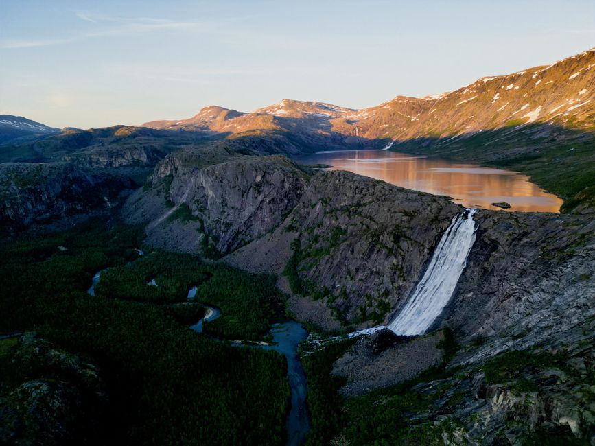A stunning aerial view of a waterfall cascading down a cliff with mountains and a lake in Norway.